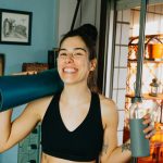 Cheerful woman with yoga mat and bottle ready for home workout in cozy room.