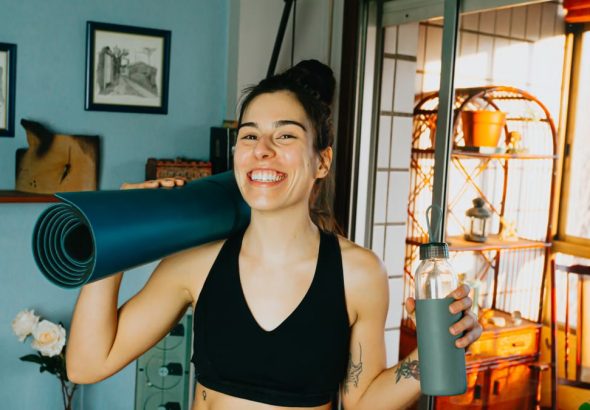 Cheerful woman with yoga mat and bottle ready for home workout in cozy room.