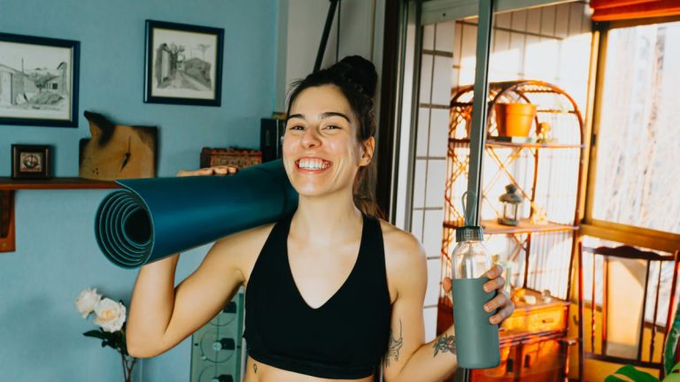 Cheerful woman with yoga mat and bottle ready for home workout in cozy room.